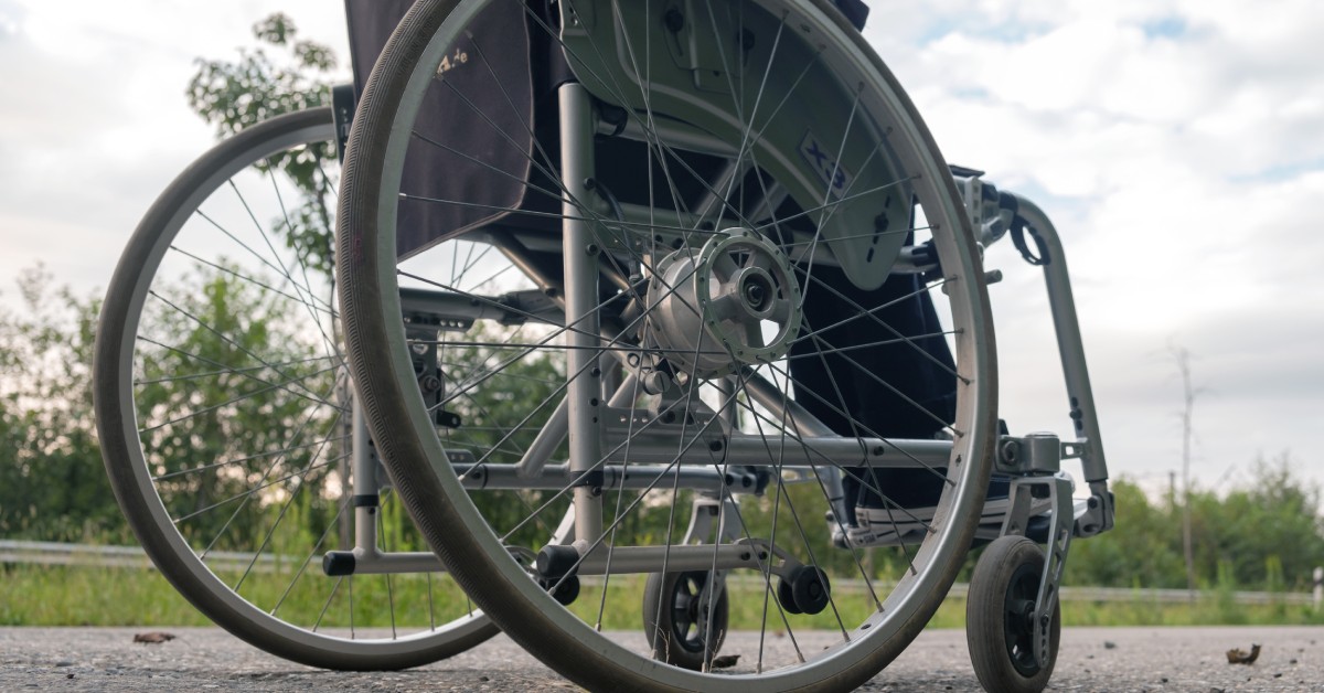 A close up of a wheelchair's tires on gray asphalt with green grass and a cloudy sky in the background.