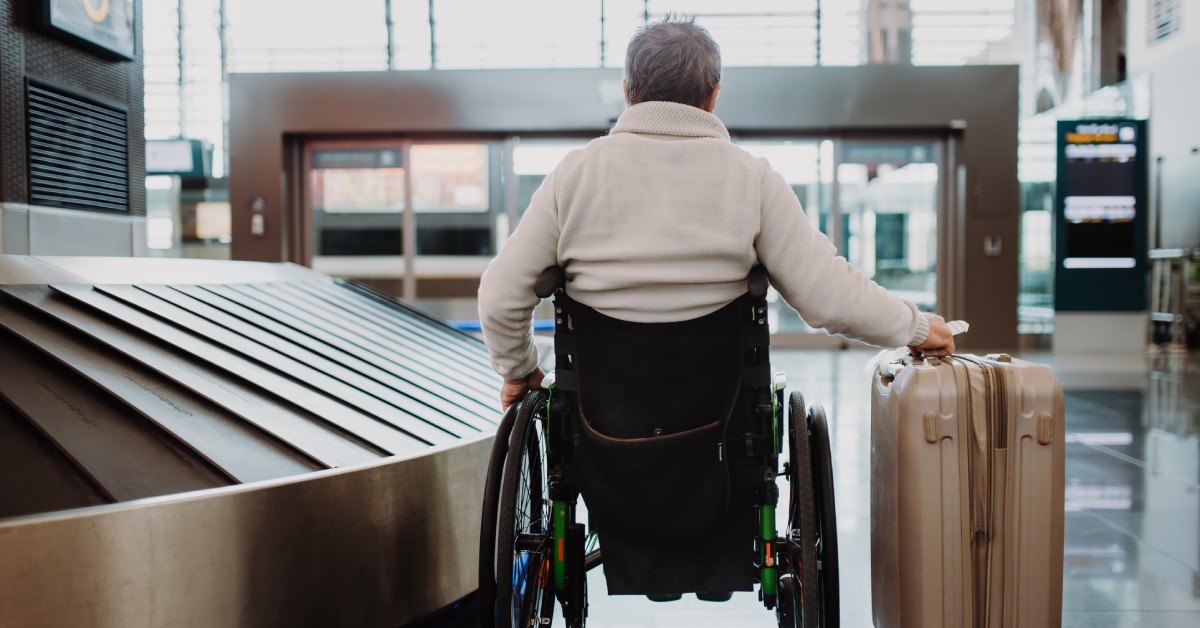 A rear view of a man sitting in his wheelchair inside of an airport with the luggage claim on his left. His luggage is to his right.