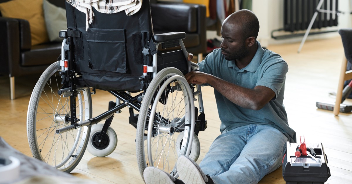 A man sits on the floor next to his wheelchair and a tool kit. The man checks his wheelchair for maintenance.