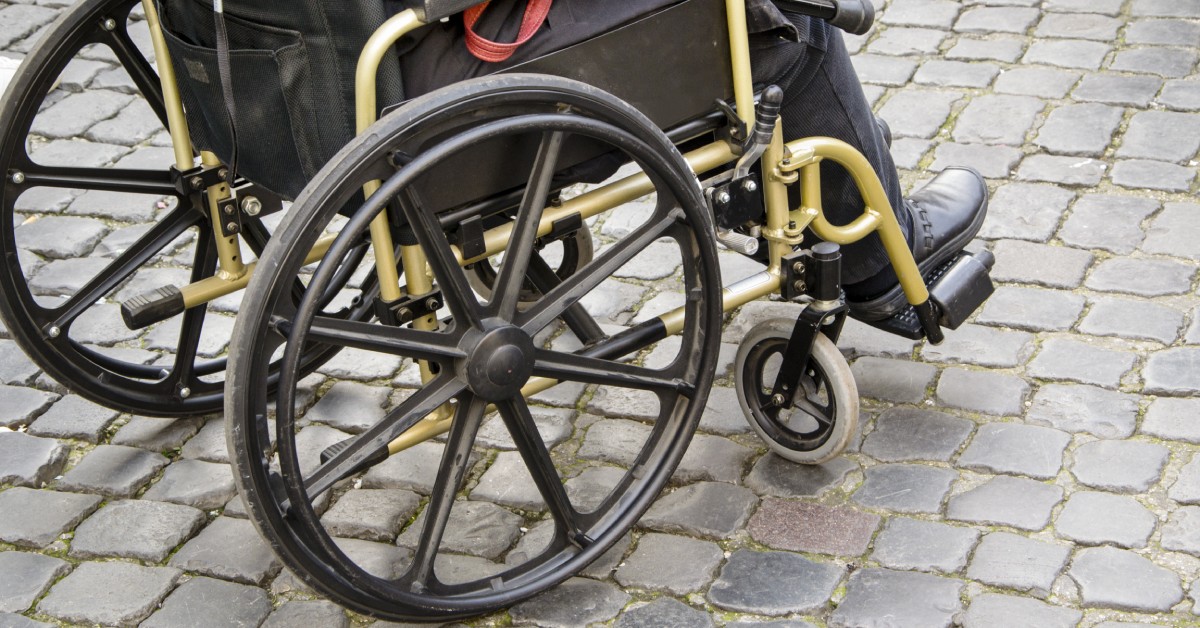 Someone sits in a wheelchair on a cobblestone street. The wheels and casters of the wheelchair are on display.