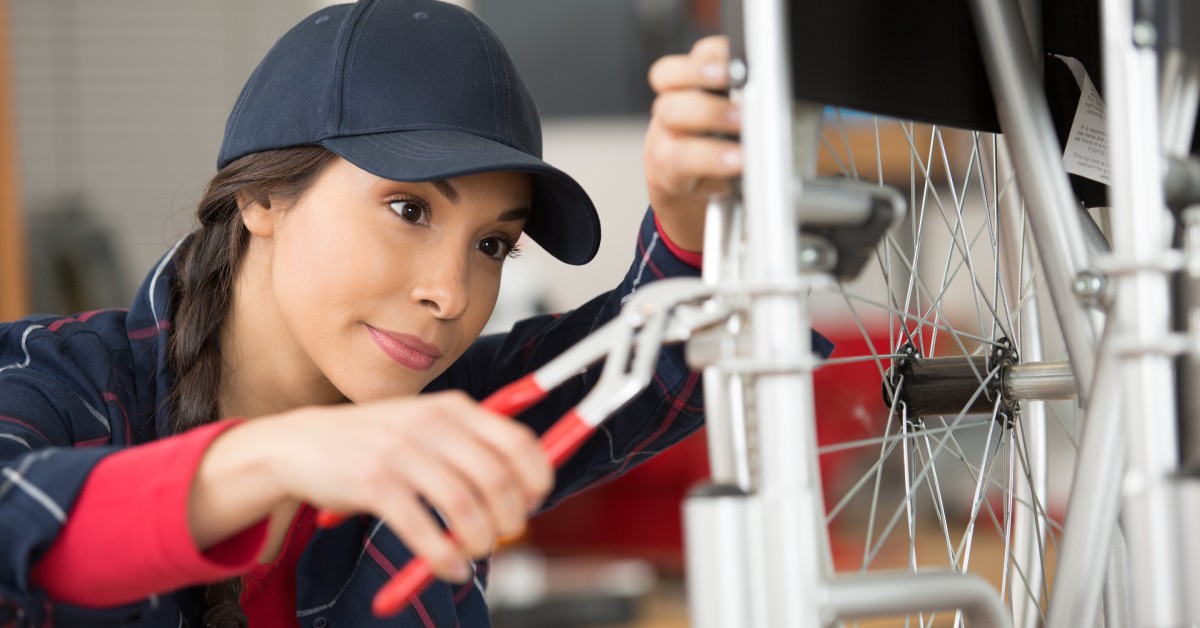 A woman wearing a black ball cap crouches next to a wheelchair. She is holding a pliers and servicing the wheelchair tire.