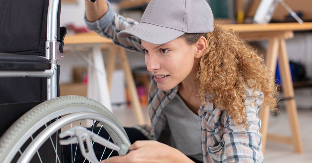 A woman is kneeling next to a wheelchair and holding a tool next to the wheel. She is wearing a grey baseball cap.