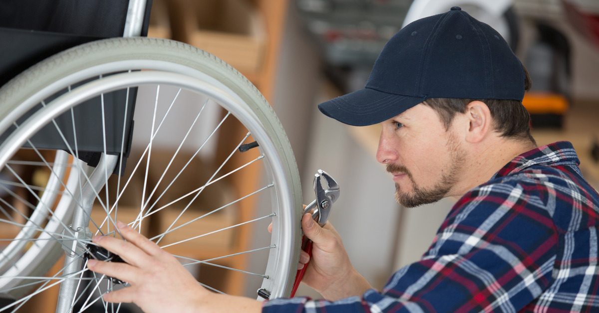 A man checks the tire of a wheelchair. He is wearing a black ballcap, a plaid shirt, and holding a tool in his right hand.