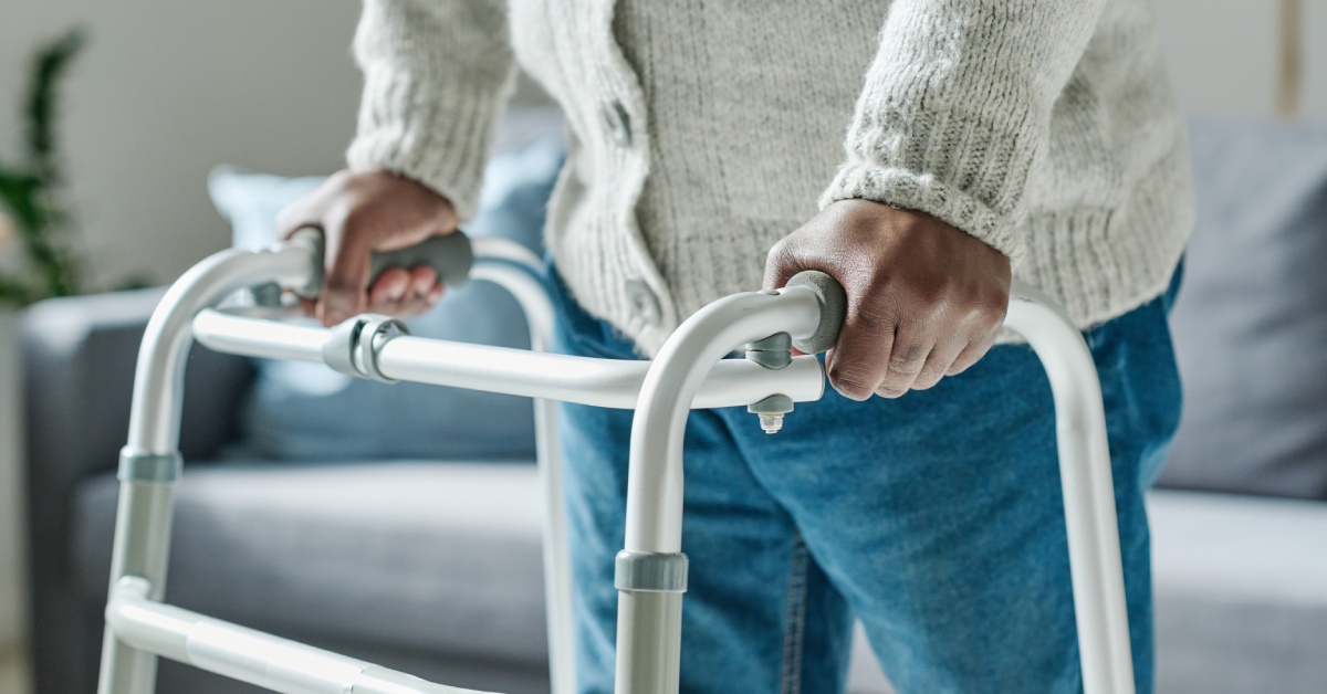 A close-up of someone using a walker to move. Their hands rest on the device, with a couch in the background.