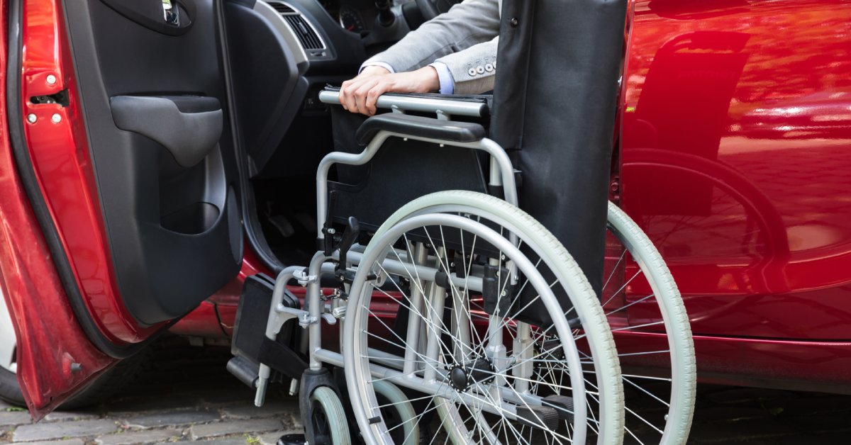 A person sits in a red vehicle with the driver's door open. They are reaching for a foldable wheelchair next to the car.