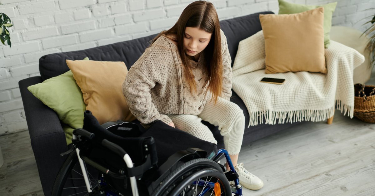 A young woman sits on her couch next to her wheelchair. One hand is on the chair like she is transferring to sit in it.