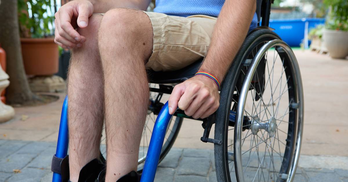 A man wearing a blue shirt and cargo shorts sitting in a blue wheelchair, holding onto the brake on the left wheel.