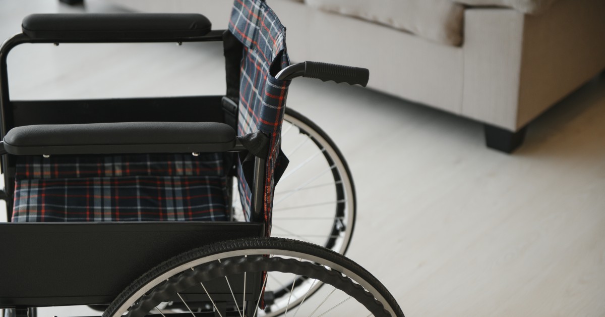 A close-up of an empty wheelchair in a living room. You can see there is a plaid cover on the seat of the wheelchair.